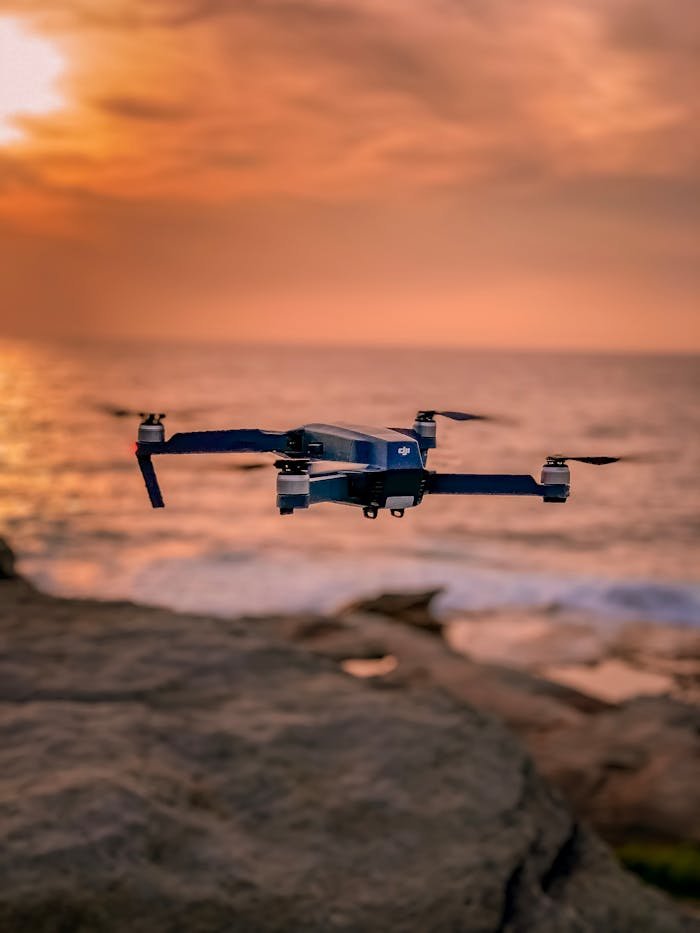 A drone hovers over a rocky shoreline with a stunning sunset backdrop, creating a serene atmosphere.